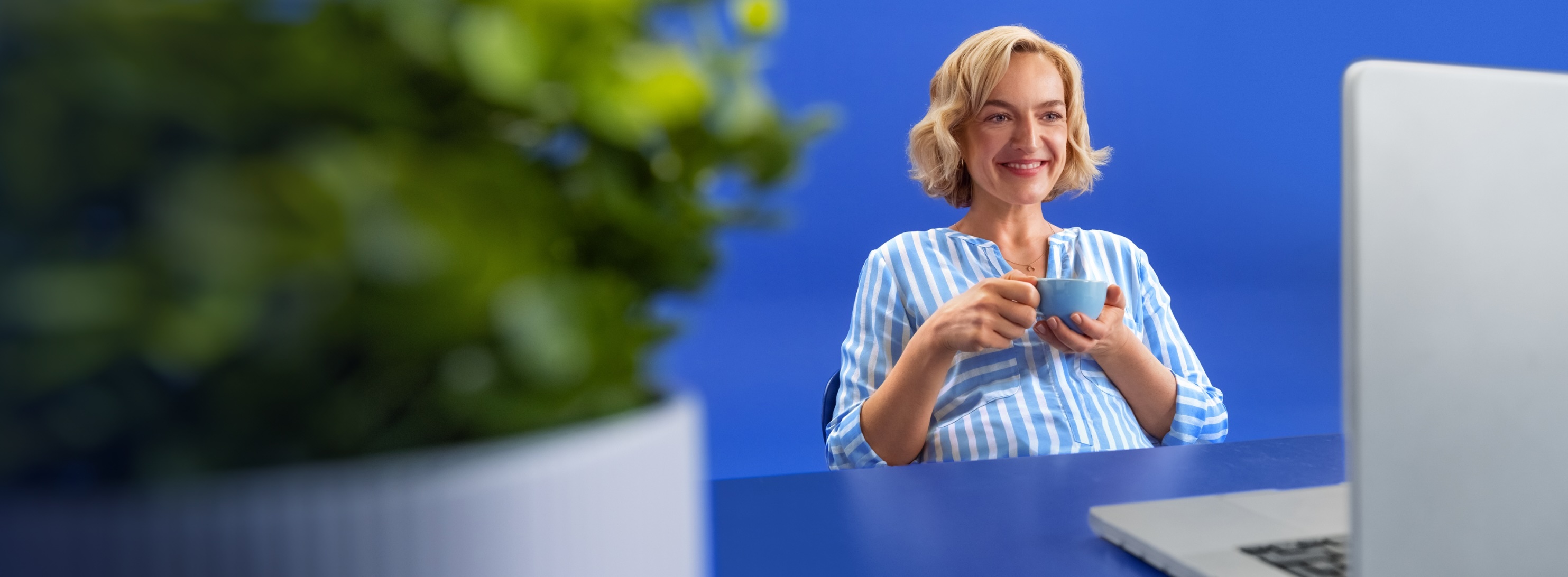 A smiling blonde woman in a blue striped top is sitting at a blue desk, in front of a blue background.  In front of her is a laptop and she is holding a cup in her hands. On the left of the picture is a plant.